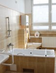 Łazienka dla mieszkańców. Na ścianach kremowe płytki ceramiczne. W centralnej części wanna. W tle okno przesłonięte poziomymi żaluzjami